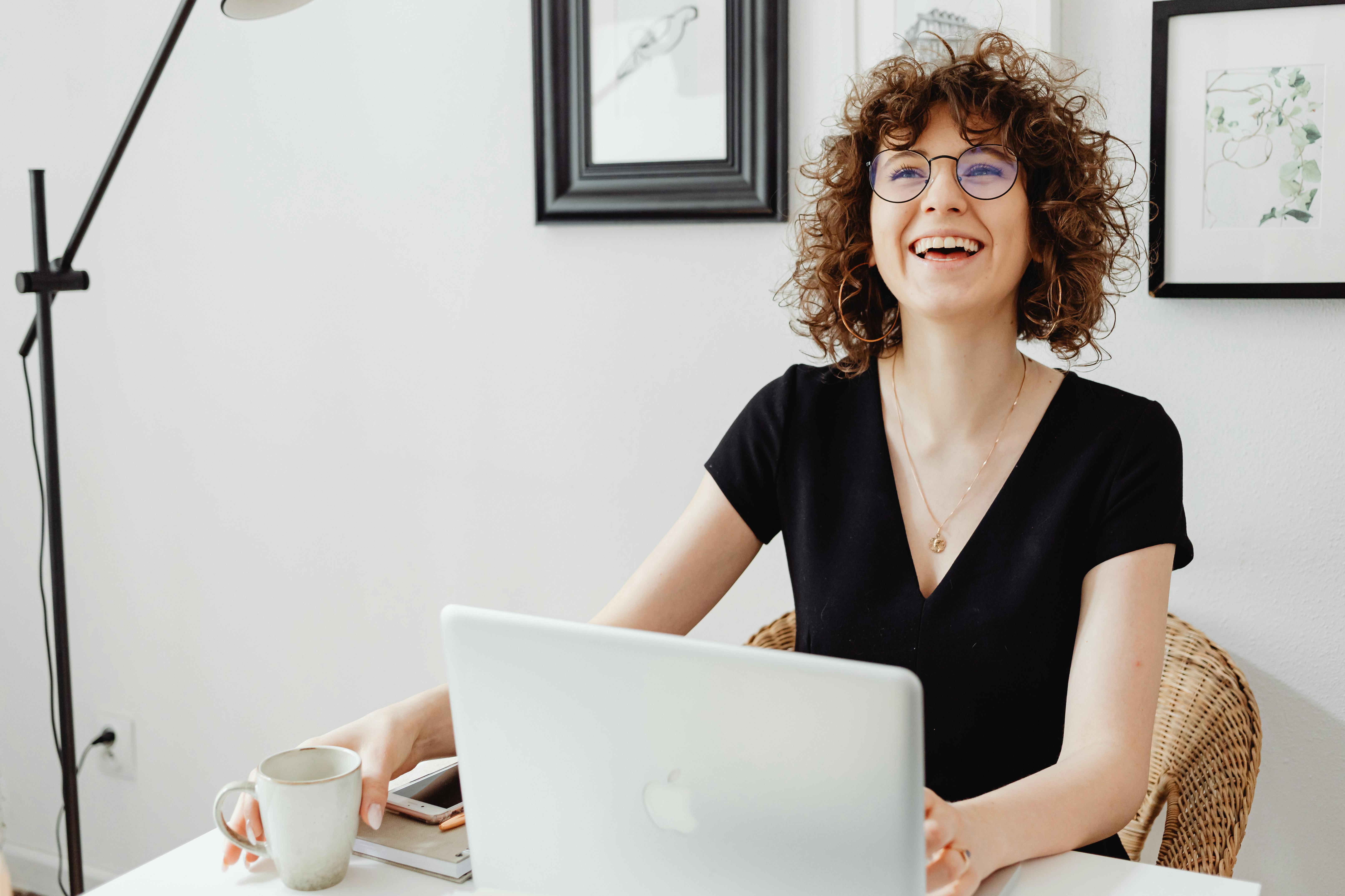 Une jeune femme souriante travaille sur un ordinateur portable blanc dans un cadre intérieur lumineux, tasse à café à la main, ambiance agréable et détendue.